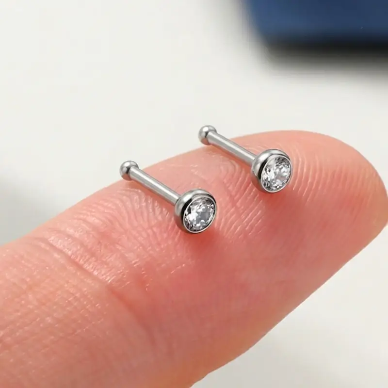 Two silver-tone bone nose studs resting on a fingertip, showing their small size